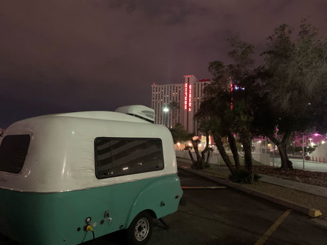 Night view of a retro **rv rental** camper, white & turquoise, parked at Circus Circus Las Vegas. Bright neon sign and trees visible.