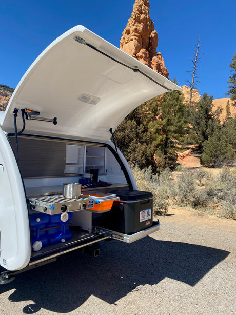 White teardrop trailer RV rental with its open rear hatch revealing a slide-out outdoor kitchen with cooktop, set against red rocks.