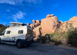 An RV rental camper van with a pop-top is parked beside majestic desert rocks under a vivid blue sky, ready for scenic adventures.
