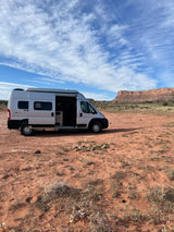 A white Winnebago Solis rv rental parked on red desert earth with an open side door, facing a majestic mesa under blue skies.