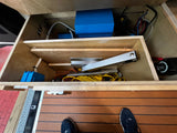 RV rental utility bay: blue electrical devices, wires, and storage organized within a wooden cabinet. Person's shoe visible on the floor.
