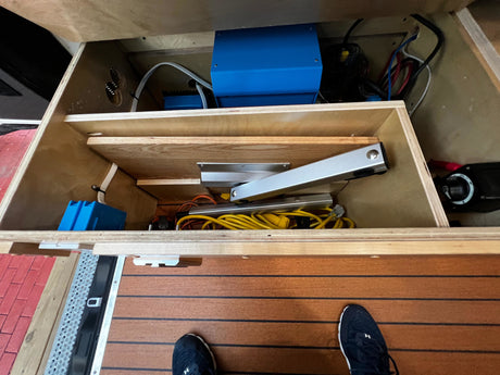 RV rental utility bay: blue electrical devices, wires, and storage organized within a wooden cabinet. Person's shoe visible on the floor.