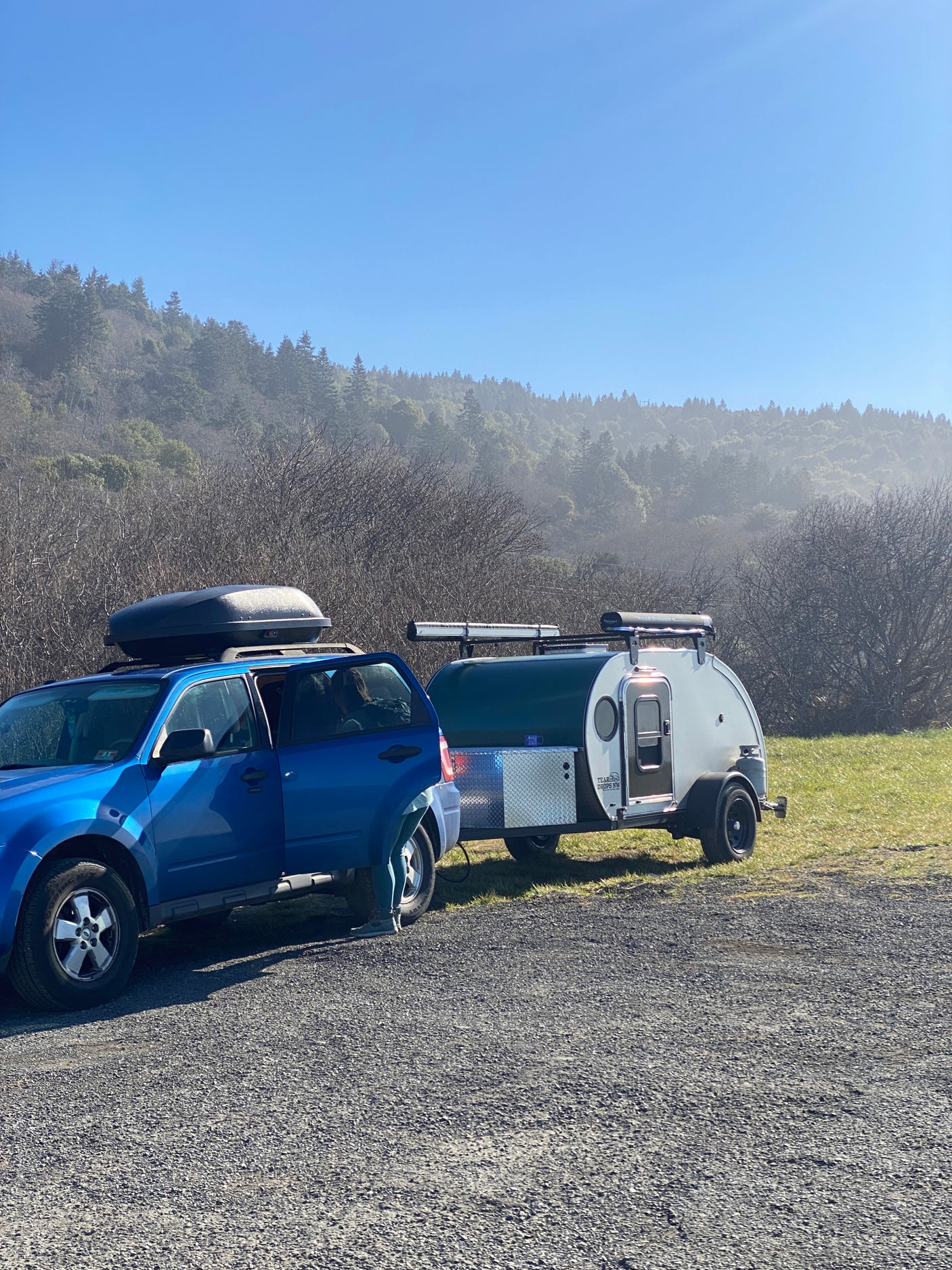 Blue SUV with roof box towing a compact teardrop RV rental. Parked in a gravel lot with misty forest background.