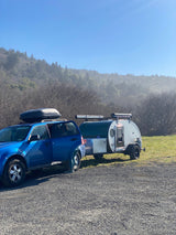 Blue SUV with roof box towing a compact teardrop RV rental. Parked in a gravel lot with misty forest background.