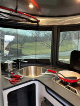Modern RV rental kitchen with sink, stovetop, and red cookware. Large windows offer a view of a grassy, treed landscape.