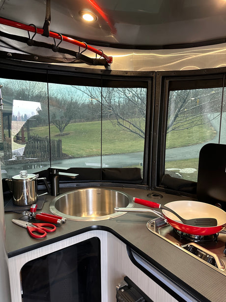Modern RV rental kitchen with sink, stovetop, and red cookware. Large windows offer a view of a grassy, treed landscape.