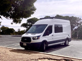 White Ford Transit RV rental camper van with roof rack, parked in a paved, tree-lined lot.