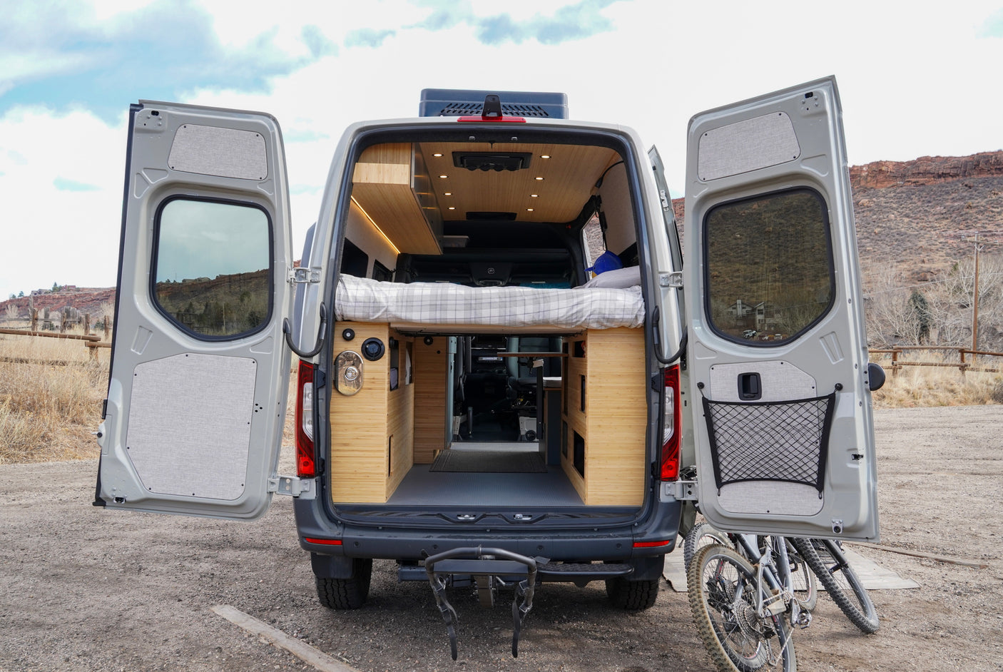 Inside a spacious rv rental camper van with open back doors. It features a custom wood interior, elevated bed, and rear bike rack.
