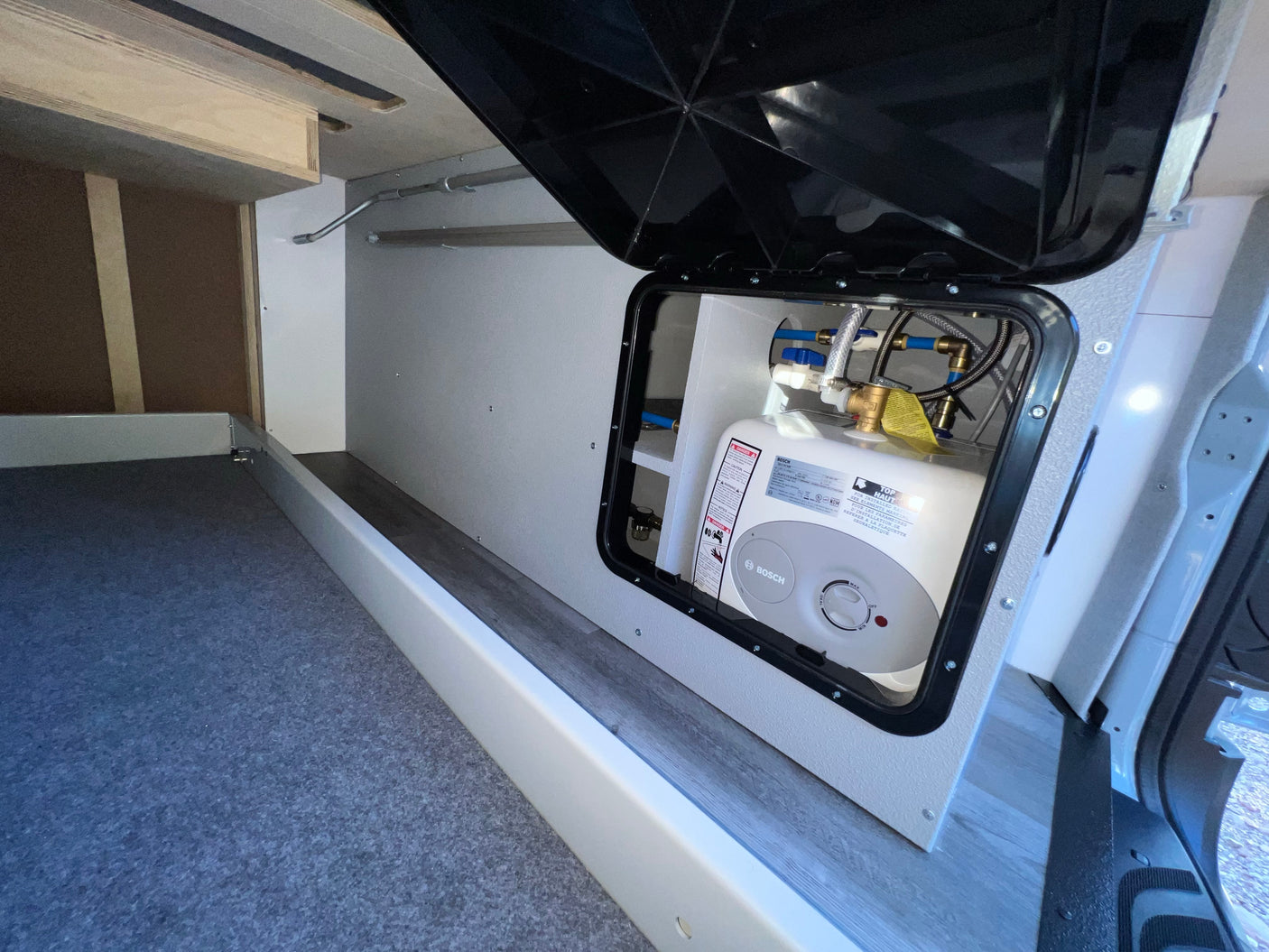 RV rental: An open access panel reveals a Bosch water heater and its plumbing, integrated into the clean camper interior with storage.