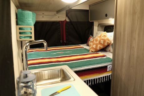 Inside a cozy rv rental: a vibrant bed with Mexican blanket, compact kitchen sink, & upper storage for camping.