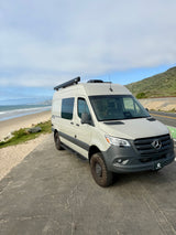 Light-tan Mercedes Sprinter rv rental van with off-road tires parked by a sandy beach, ocean, and hills. Ready for adventure.
