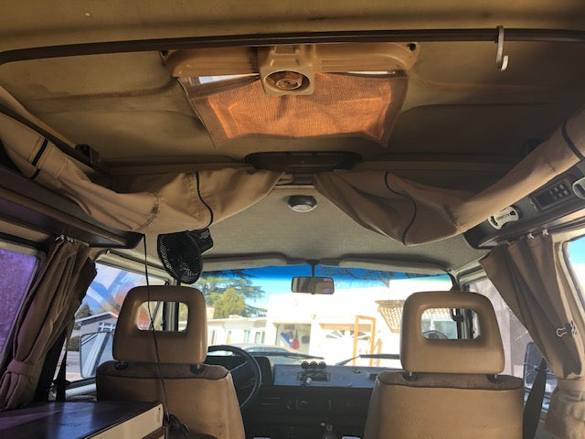 This RV rental's interior reveals the front seats, dashboard, overhead storage, and a small fan. A clear view out the windshield.