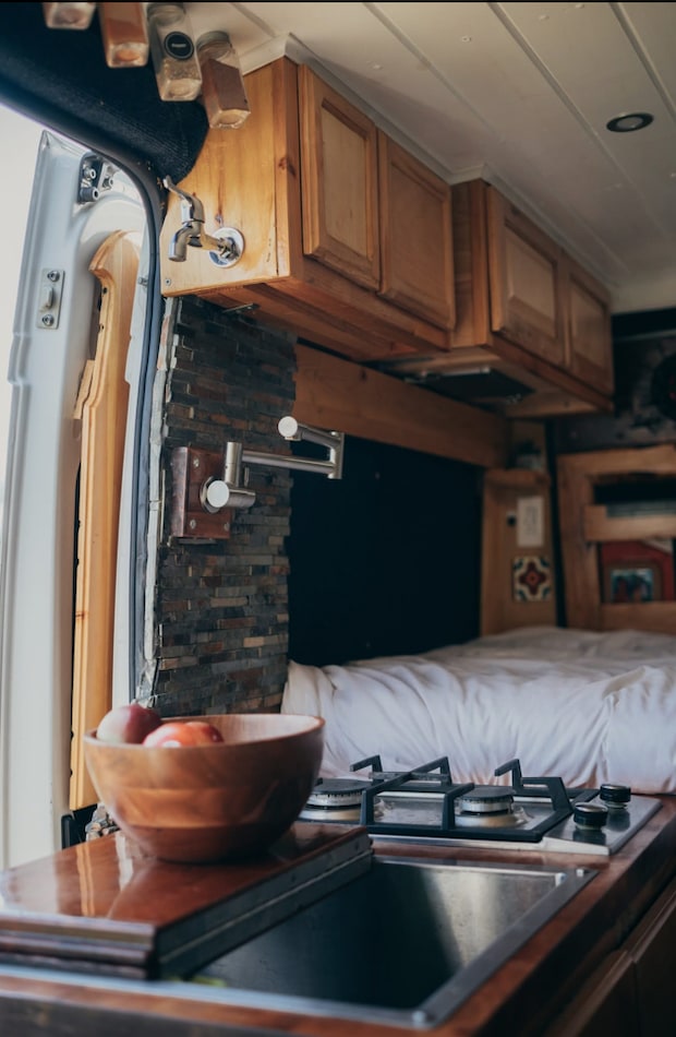 Explore your RV rental's interior: a compact kitchen with stove, sink, and wood cabinets, next to a cozy bed.