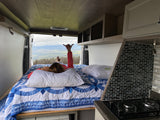 Cozy RV rental interior: A person relaxes on a bed, enjoying panoramic mountain views from open back doors. Features a kitchenette.