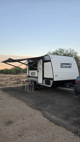 RV rental: A Coleman travel trailer with awning extended, table & chair outside, parked in a desert setting.