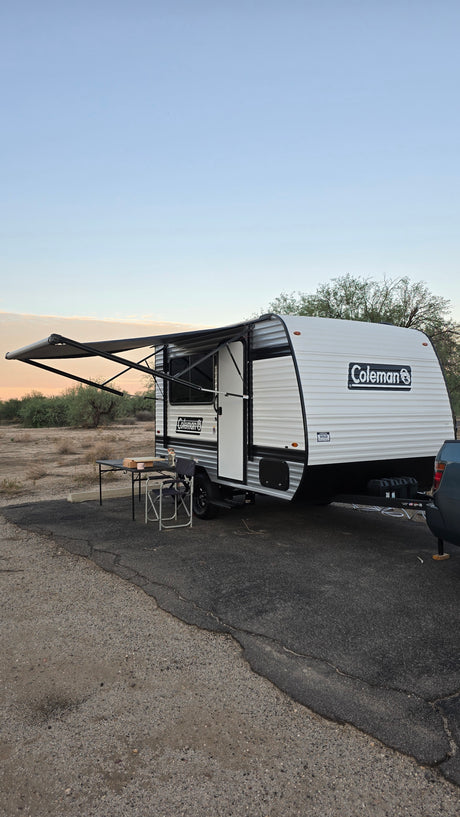 RV rental: A Coleman travel trailer with awning extended, table & chair outside, parked in a desert setting.