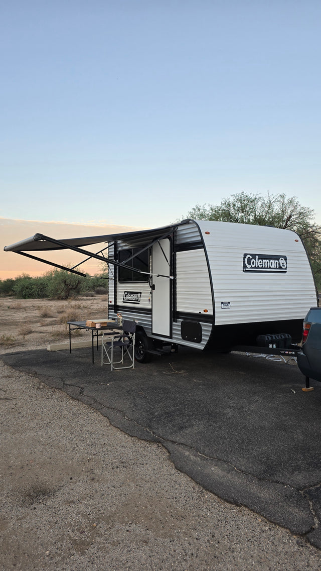 RV rental: A Coleman travel trailer with awning extended, table & chair outside, parked in a desert setting.