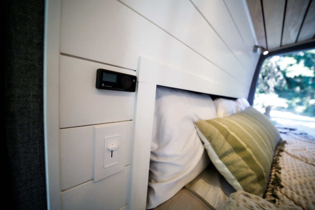 Inside a cozy RV rental: a bed with pillows, a digital heater control, and light dimmer on white wood paneling.