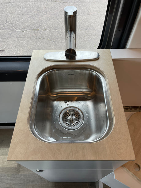 Modern sink and faucet on a wood countertop over a gray cabinet, seen inside an RV rental. Paved ground visible outside.