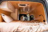 Inside a cozy RV rental van, featuring a comfortable bed, wooden walls, and overhead storage. A person stands near the front.