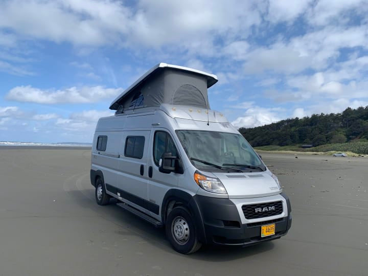 Experience van life with an rv rental! A silver camper van, pop-top open, parked on a sandy beach under a cloudy sky.