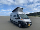 Experience van life with an rv rental! A silver camper van, pop-top open, parked on a sandy beach under a cloudy sky.
