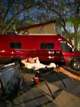 Relaxing by a red pop-top RV rental at night. A person unwinds in a chair beside the camper van setup.