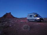 An RV rental camper van parked at dusk in a red rock desert with a large butte under a deep blue sky.