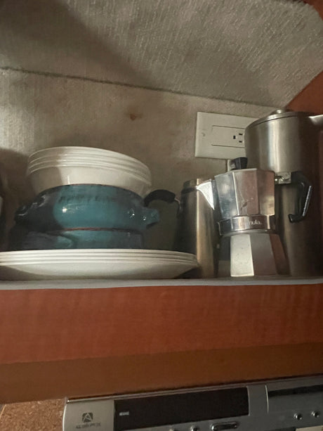 RV rental kitchen shelf: Stacked plates, a blue pot, Moka pot, and thermos, compactly stored next to an outlet.