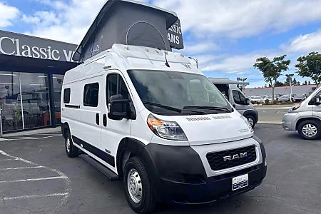White Ram ProMaster camper van with pop-top roof for RV rental, parked at Classic Vans. Other vans nearby.