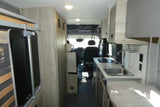 RV rental camper van interior. Features a compact kitchen with sink/stove, overhead cabinets, and a living area leading to the cockpit.