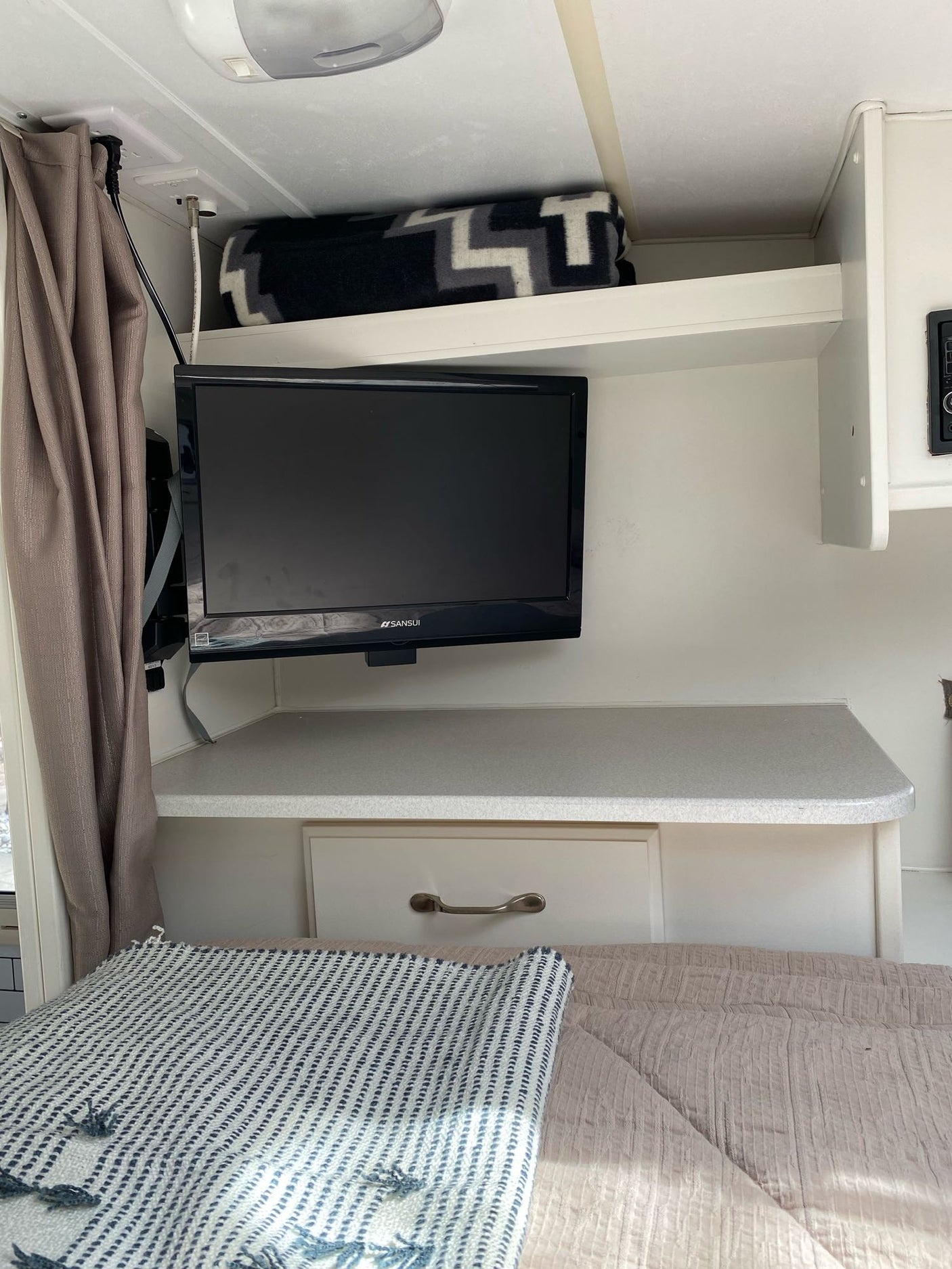 Cozy RV rental bedroom area with a wall-mounted Sansui TV, countertop, storage, and a comfy bed with blankets.