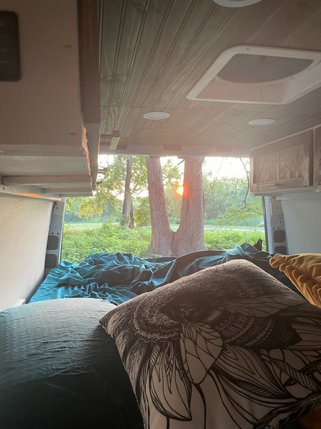 Cozy RV rental bedroom view with teal bedding, custom ceiling, and a vibrant sunrise/sunset through the trees.