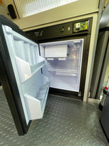 Inside an **RV rental**, a clean, empty mini-refrigerator is open. Above it, a panel displays tank levels and battery condition.