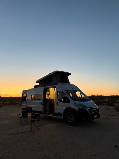 An RV rental camper van is parked in the desert at sunset, its interior glowing warmly. Chairs invite a cozy evening.