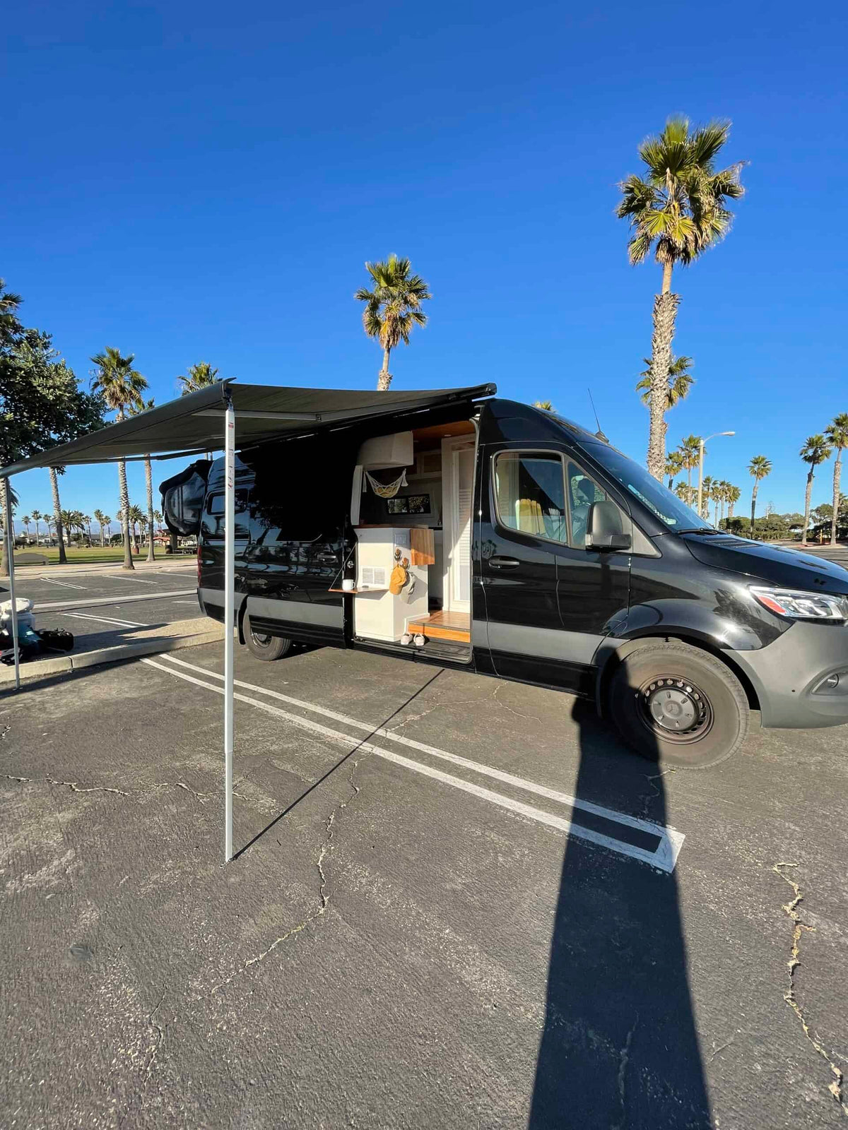 Black RV rental van with extended awning. Interior visible as it's parked by palm trees on a sunny day.|Experience comfort in this RV rental! Features a cozy bed