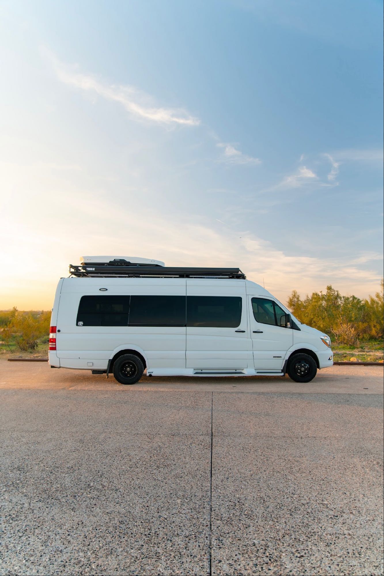 A white Mercedes-Benz Sprinter camper van with a roof rack, parked on asphalt at sunset. Ideal RV rental for your adventure.