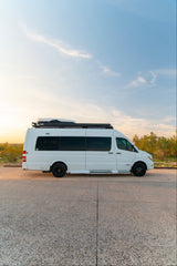 A white Mercedes-Benz Sprinter camper van with a roof rack, parked on asphalt at sunset. Ideal RV rental for your adventure.