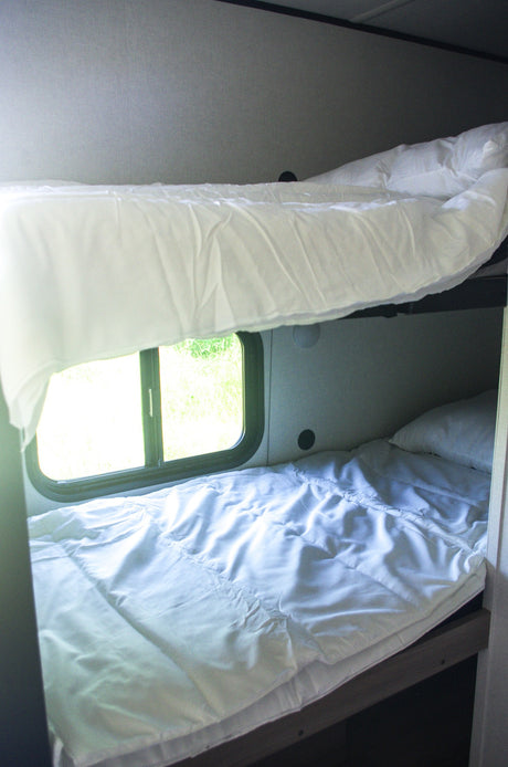 Clean, cozy bunk beds with white linens in an RV rental. A window reveals green nature outside, ideal for travel.