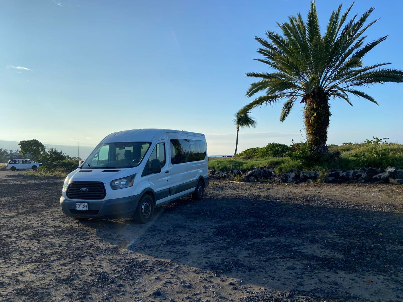 A white Ford Transit rv rental van on a gravel lot under clear skies. Palm trees and distant ocean add to this scenic travel setting.