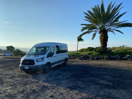 A white Ford Transit rv rental van on a gravel lot under clear skies. Palm trees and distant ocean add to this scenic travel setting.