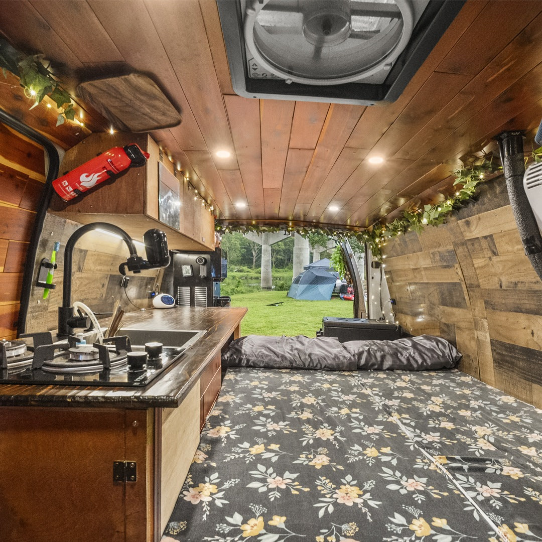 Cozy wood-paneled RV rental interior with kitchen, floral bed, and open back revealing a grassy campsite with tents.