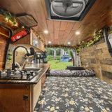 Cozy wood-paneled RV rental interior with kitchen, floral bed, and open back revealing a grassy campsite with tents.