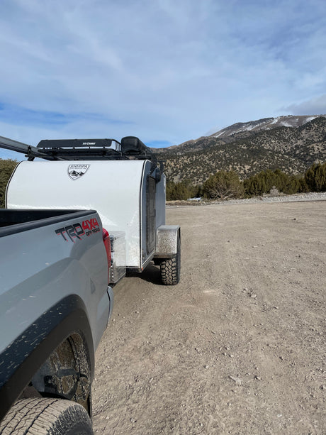 Overland ready! A grey truck tows a Sherpa teardrop trailer on a dirt road by mountains. Great RV rental for adventure.