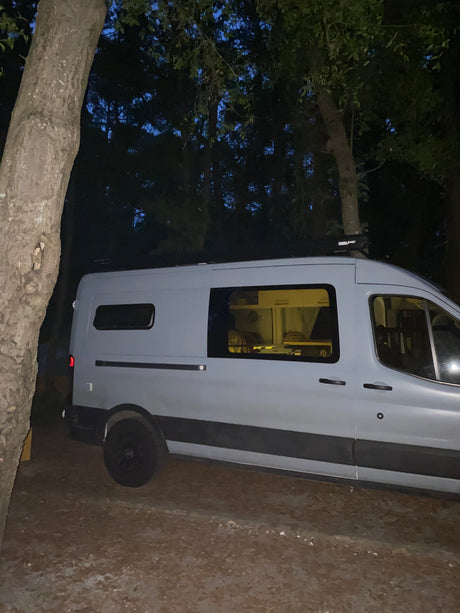 Night scene: A light gray camper van, possibly an RV rental, parked amidst trees with its interior lights on, offering a cozy retreat.