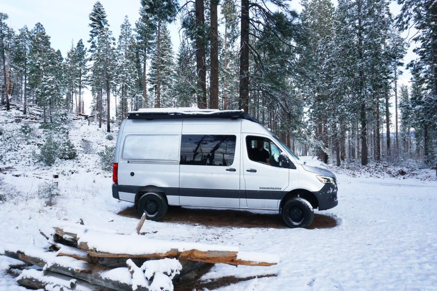 Silver Rossmonster camper van RV rental parked in a snowy winter forest, surrounded by tall snow-laden pine trees.