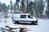 Silver Rossmonster camper van RV rental parked in a snowy winter forest, surrounded by tall snow-laden pine trees.