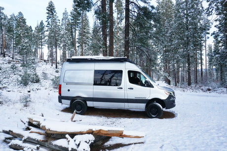 Silver Rossmonster camper van RV rental parked in a snowy winter forest, surrounded by tall snow-laden pine trees.