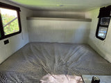 Inside an RV rental: a compact sleeping area with a checkered bed, overhead shelf, and windows showing nature & a Jeep.