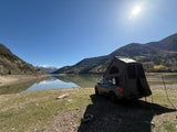 A truck with a rooftop tent is set up for camping beside a calm mountain lake, an adventurous **RV rental** alternative.